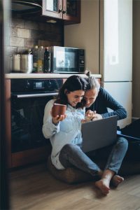 Couple sitting on kitchen floor, using a laptop to explore mortgage calculator for refinancing options, with a coffee mug in hand.