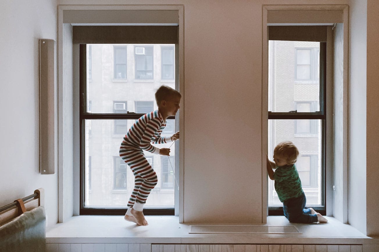 Two children playing in a bright room, one jumping in striped pajamas and the other kneeling by a window, emphasizing a playful home environment.