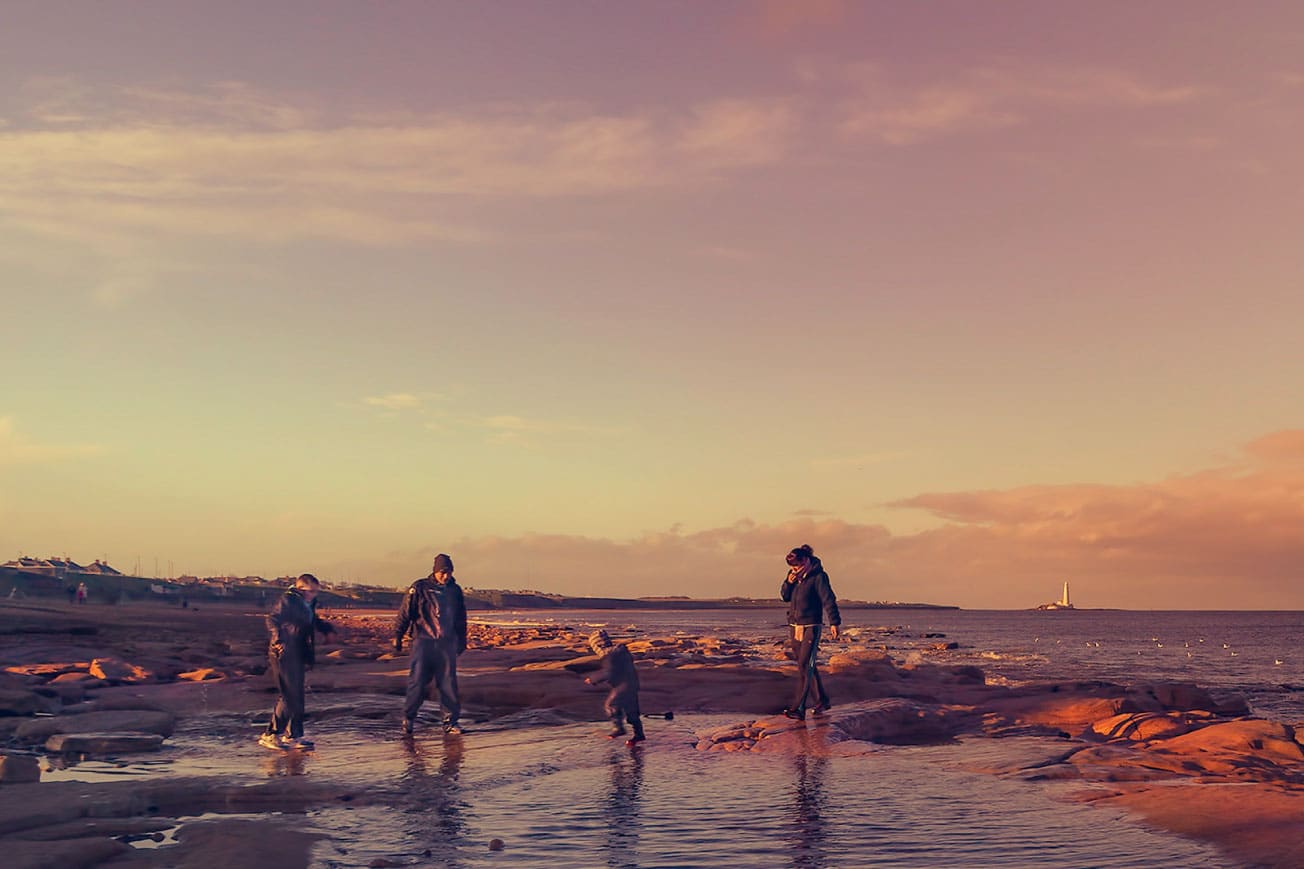Family exploring rocky beach during sunset, with children playing in shallow water and lighthouse in the distance, highlighting outdoor living and seasonal family activities.