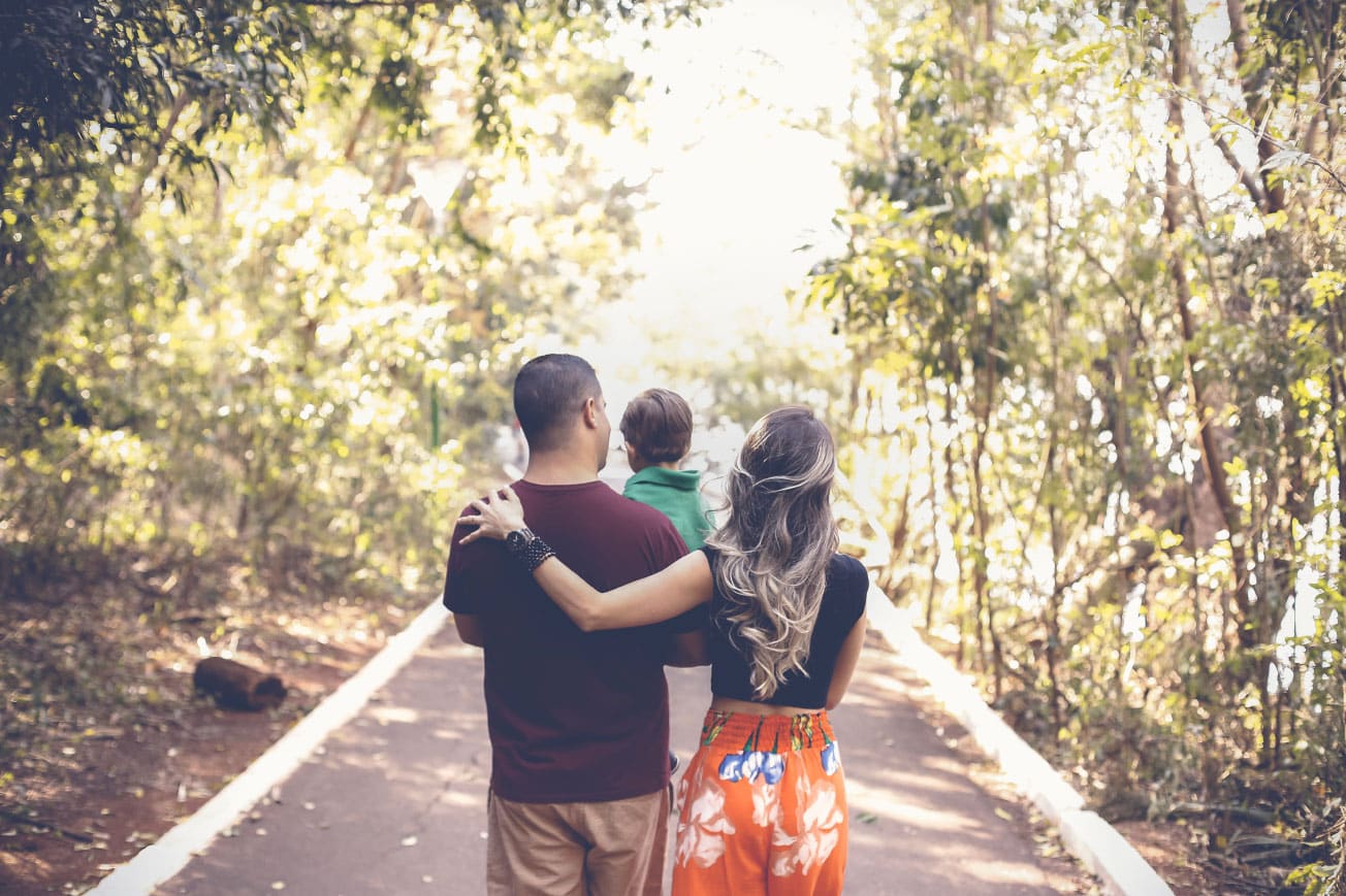 Family walking together in a lush green park, with a father holding a child on his shoulders and a mother embracing him, illustrating family bonding and outdoor activities.