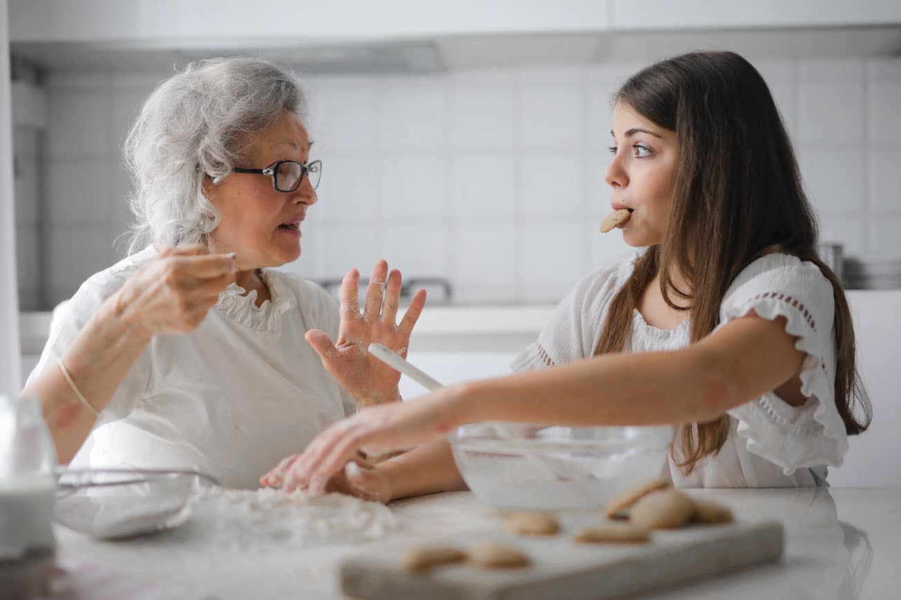 Elderly woman and young girl baking cookies together in a bright kitchen, with flour on the table and the girl playfully tasting cookie dough.