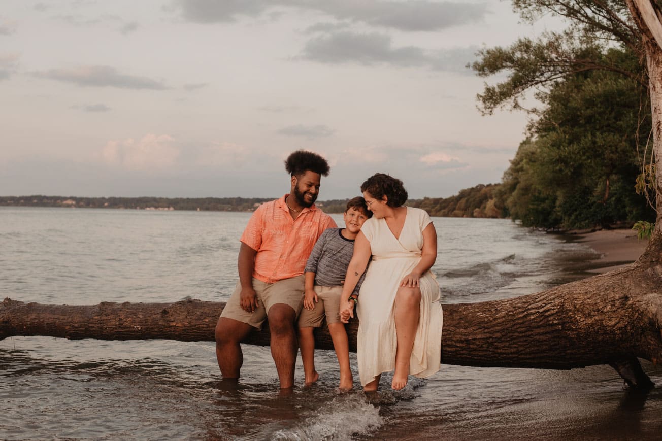Family sitting on a log by the water, enjoying a moment together, with a serene lake and trees in the background.