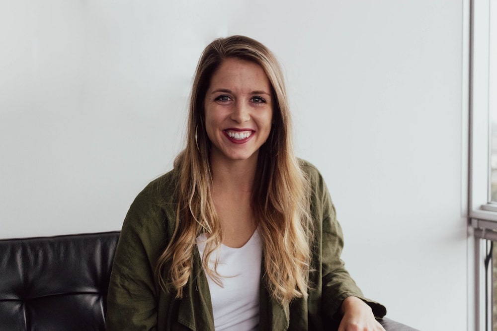 Smiling woman with long hair wearing a green jacket seated on a black couch, in a bright indoor setting.