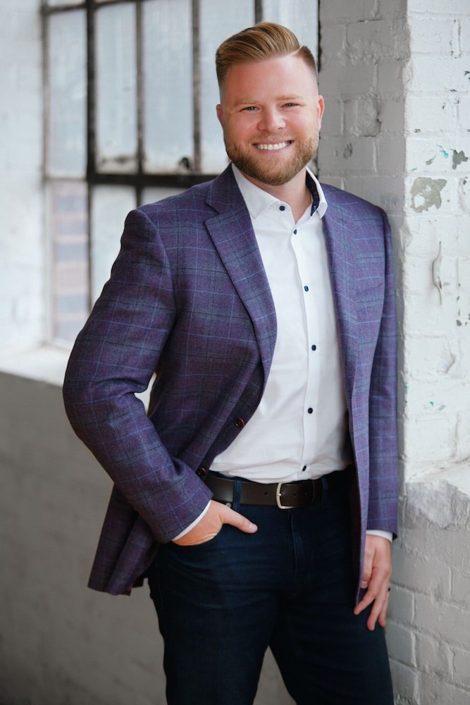 Smiling man in a purple plaid blazer and white shirt, standing against a brick wall, representing professionalism and confidence.