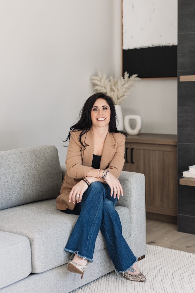 Woman sitting on a gray sofa, wearing a tan blazer and flared jeans, smiling, in a modern interior setting with decorative elements.