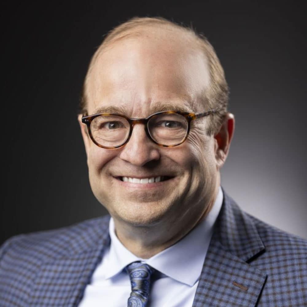 Chris Dunn, Loan Officer at Refined Lending, smiling against a dark background, wearing glasses and a checkered suit jacket, conveying professionalism and approachability.