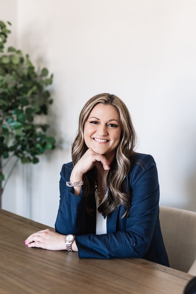 Professional woman smiling at desk, wearing navy blazer and silver jewelry, with greenery in background.