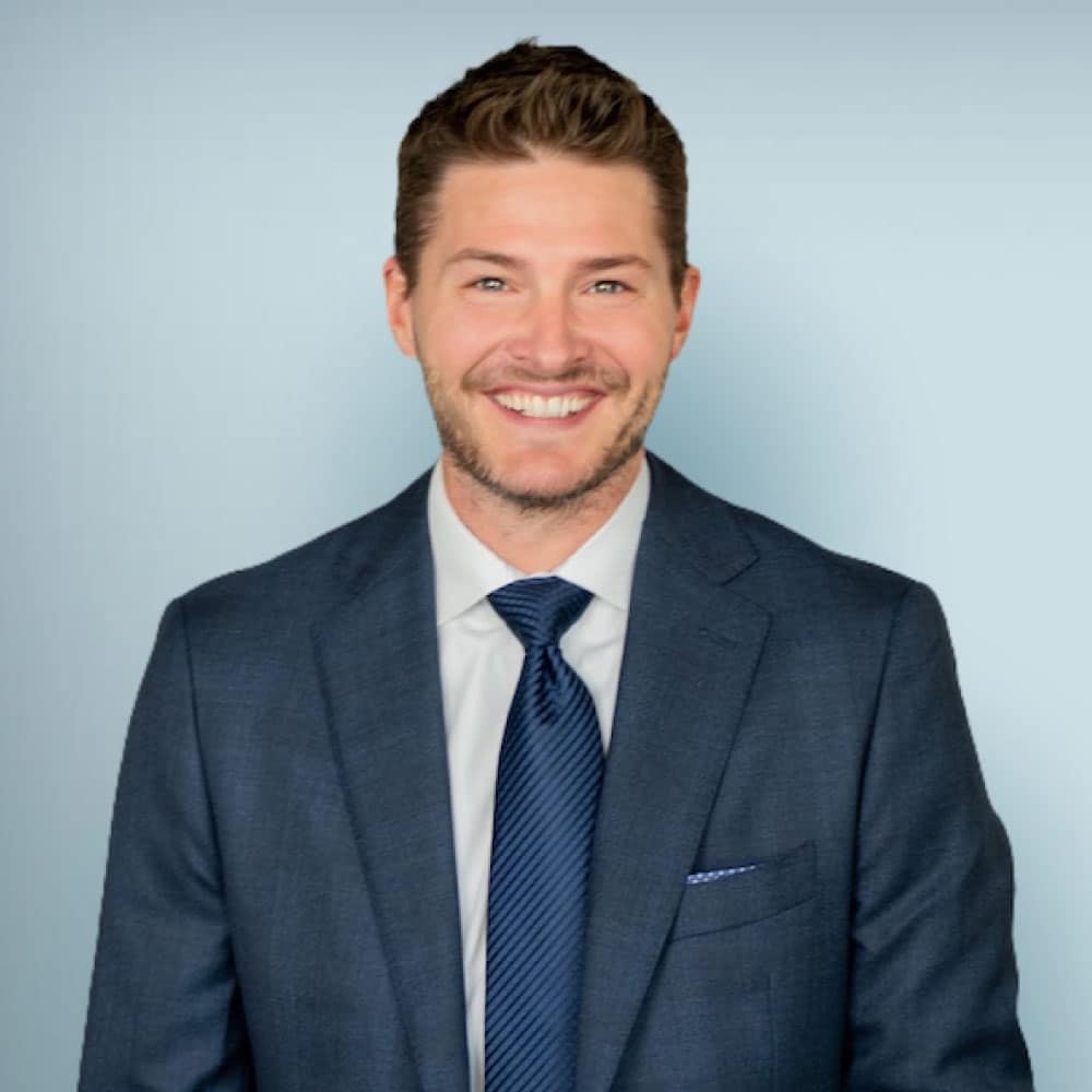 Smiling man in a professional suit, showcasing a confident demeanor against a light blue background.