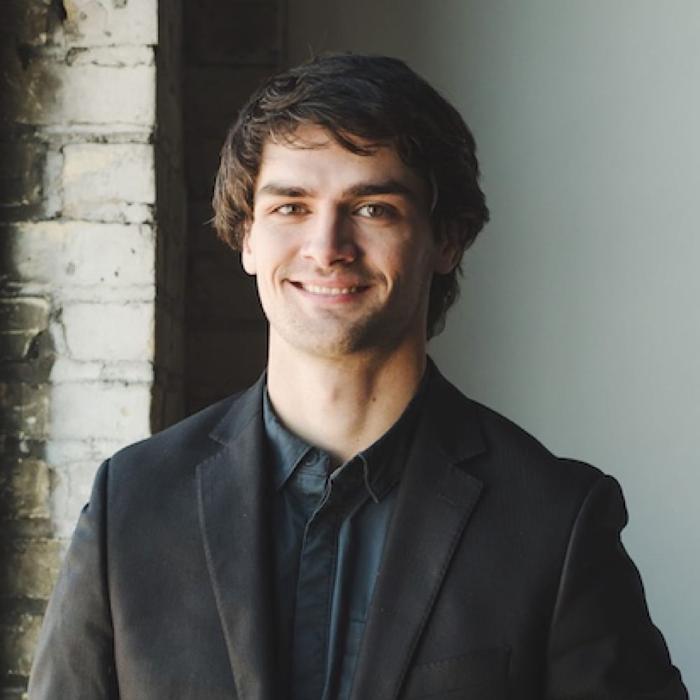 Smiling young man in a black suit standing near a brick wall, representing professional services at Refined Lending.