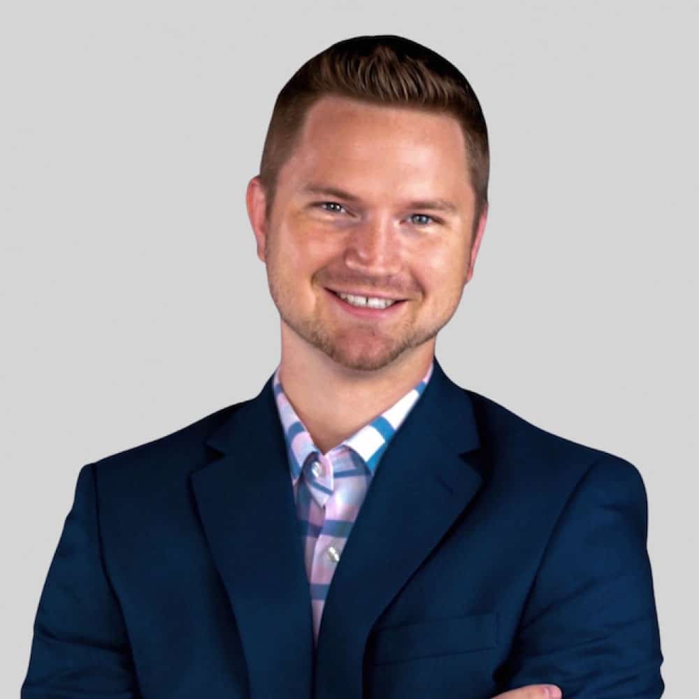 Professional man smiling in a suit with a blue jacket and patterned shirt, against a neutral gray background, representing Refined Lending.