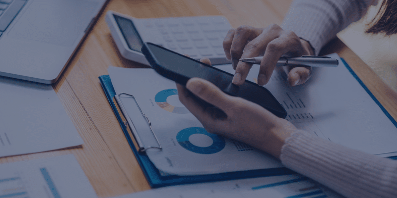 Person using smartphone and pen while analyzing financial documents with charts, calculator, and laptop, related to mortgage and refinancing options.
