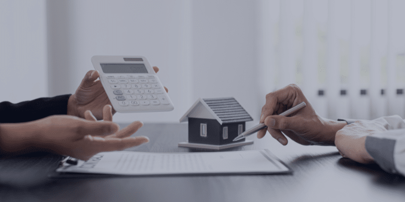 Hands discussing home financing with a calculator and a miniature house model on a table, symbolizing mortgage and home selling strategies.