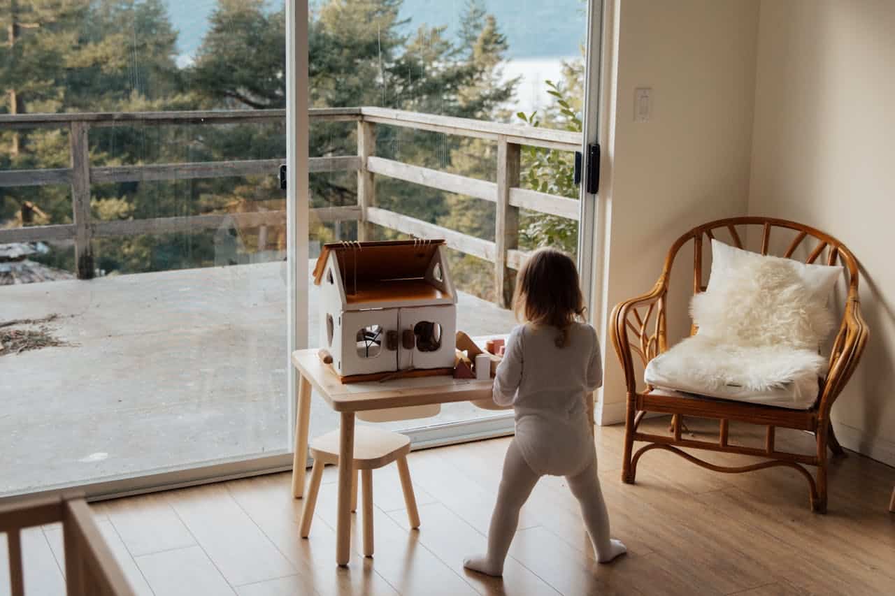 Child playing with a toy house at a table in a sunlit room, featuring large windows overlooking a natural landscape, emphasizing the importance of home and family for first-time buyers.