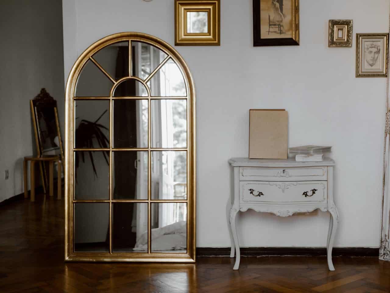 Elegant gold-framed mirror reflecting a stylish interior with a white side table and decorative art pieces on the wall, showcasing a cozy home environment.
